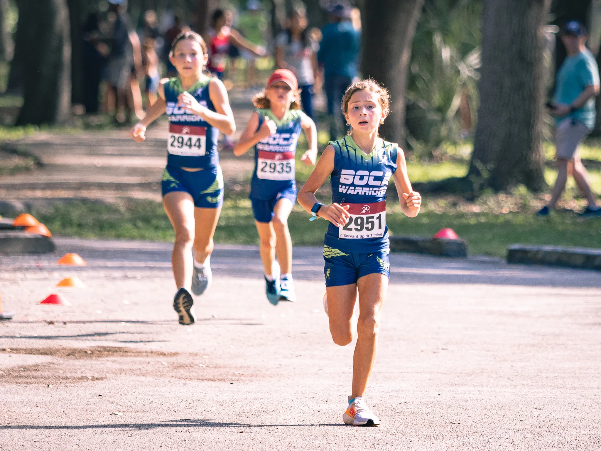 Three youth girls racing in a cross country meet on a dirt path lined with orange cones in a sunny park