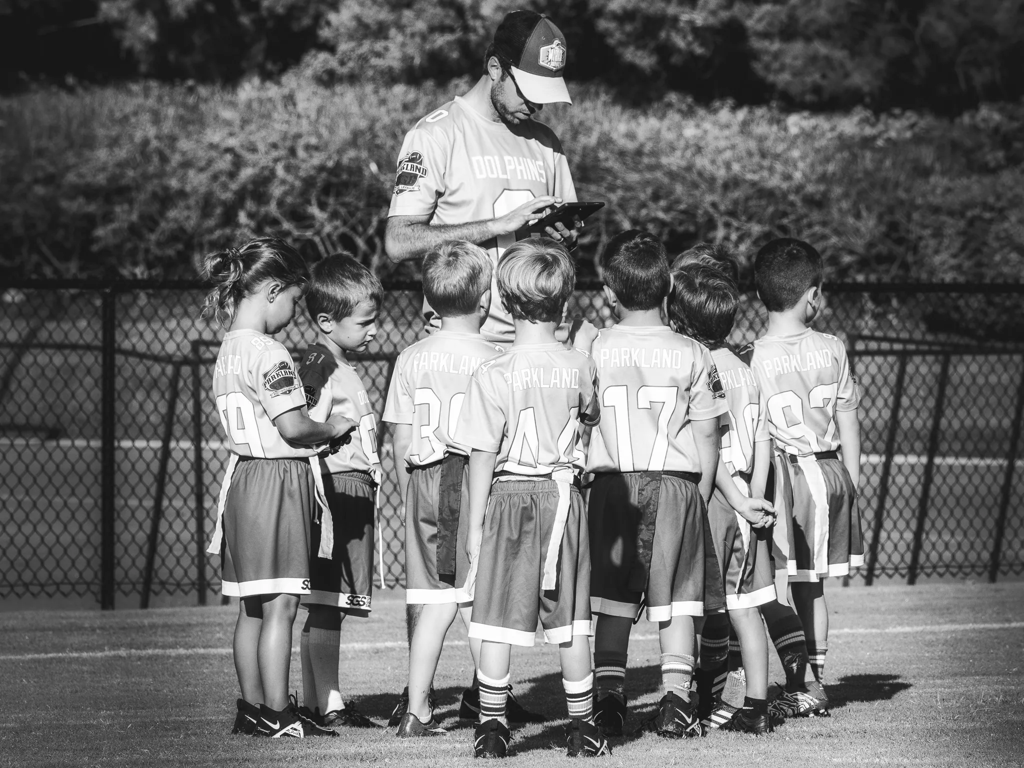 Youth flag football coach in a cap addresses a huddle of about ten young players in jerseys on a turf field, black and white