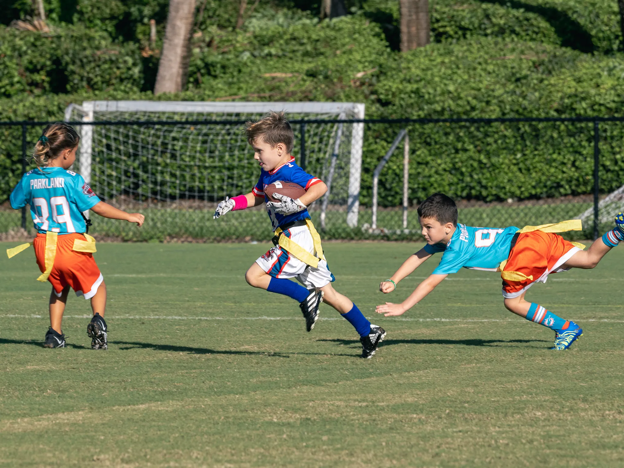 Young boy in blue flag football jersey sprints with the ball past two defenders in teal and orange on a green field
