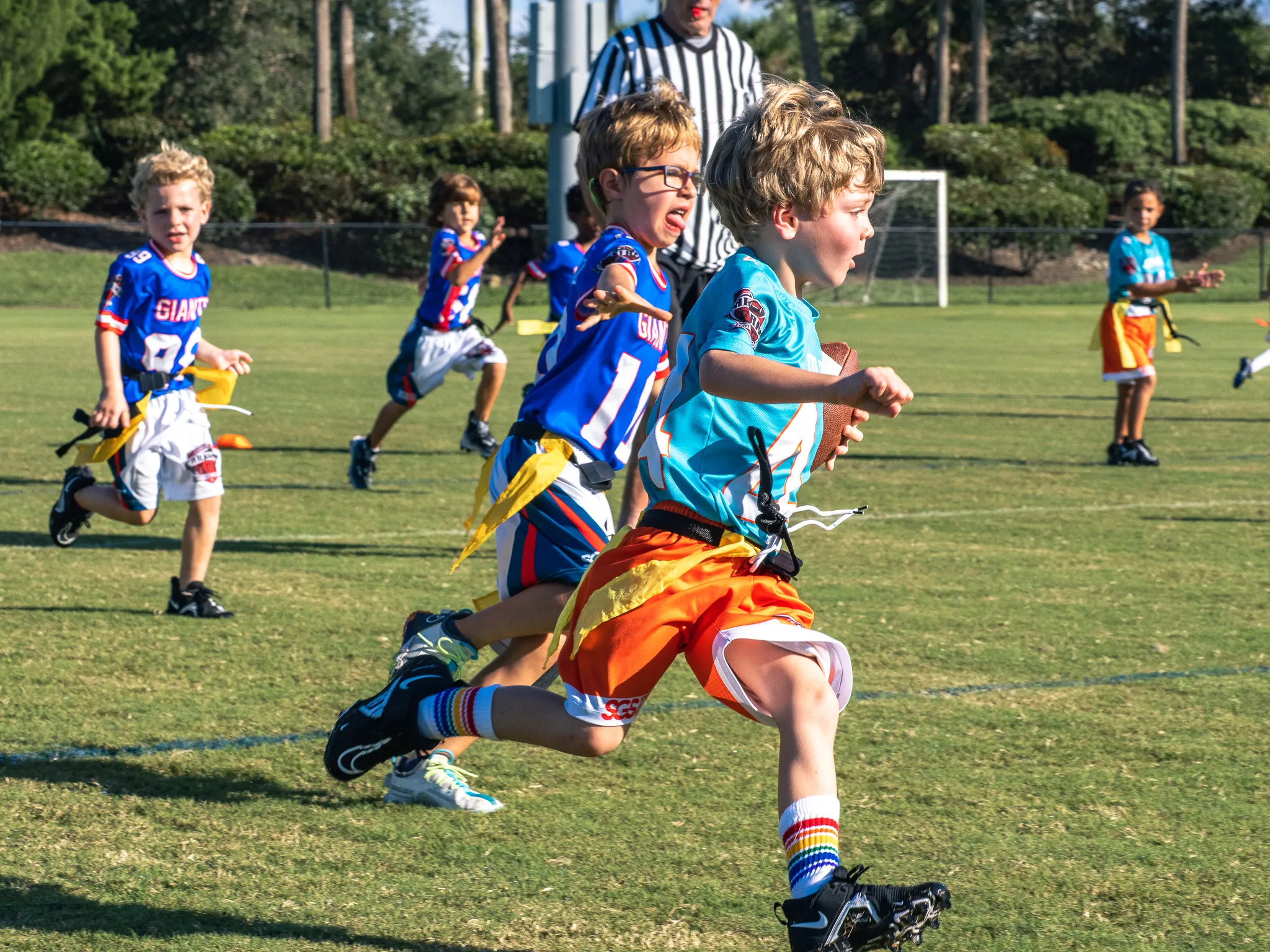 Boy in teal jersey carrying a football runs ahead of defenders during a youth flag football game on a grass field with a referee behind