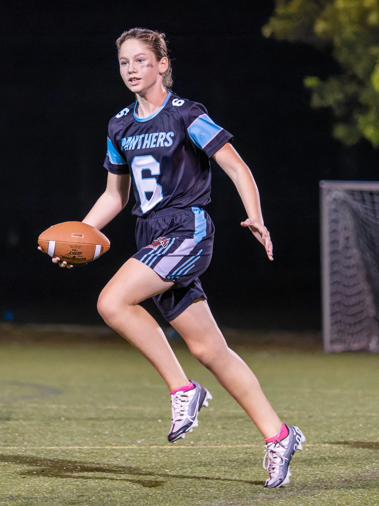 Teen girl in Panthers number 6 jersey runs with a football during a nighttime youth flag football game on a lit field