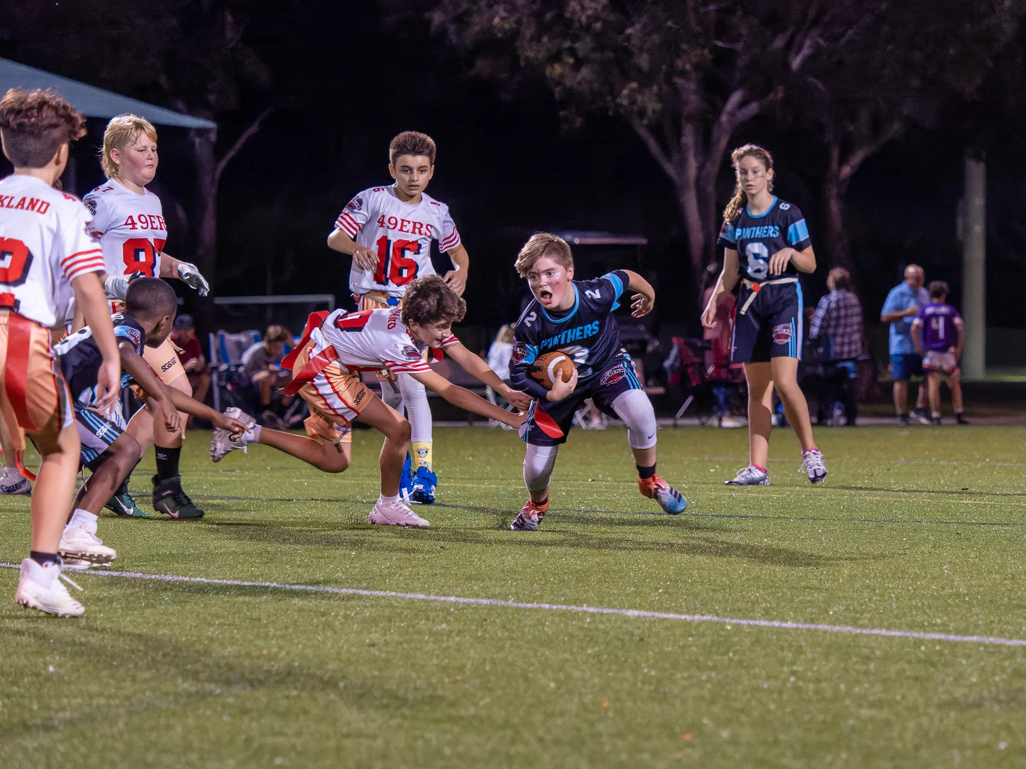 Panthers flag football player dodges a defender while running with the ball during a nighttime game with multiple teams on the field