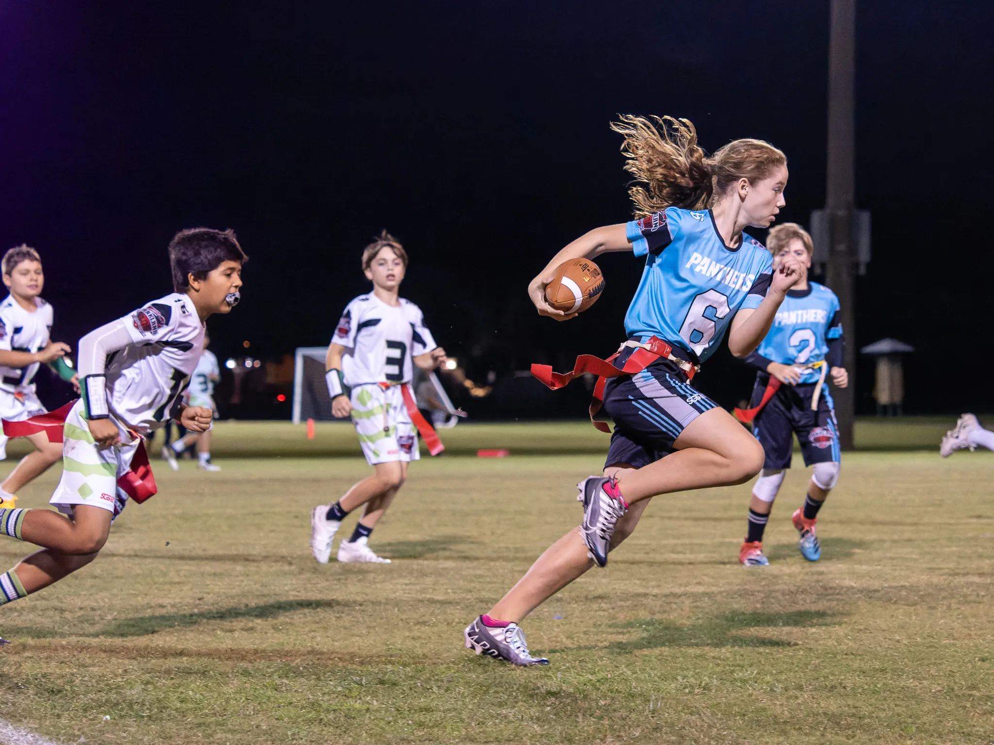 Teen girl in light blue Panthers number 6 jersey sprints with the football past defenders under field lights at night, hair flying