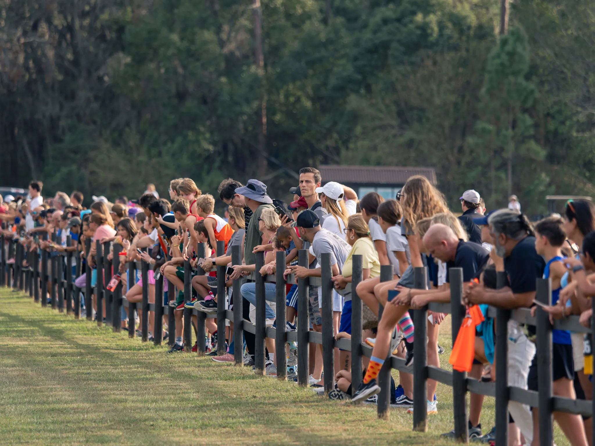 Large crowd of spectators leaning against a wooden fence watching a youth cross country race on a grassy course