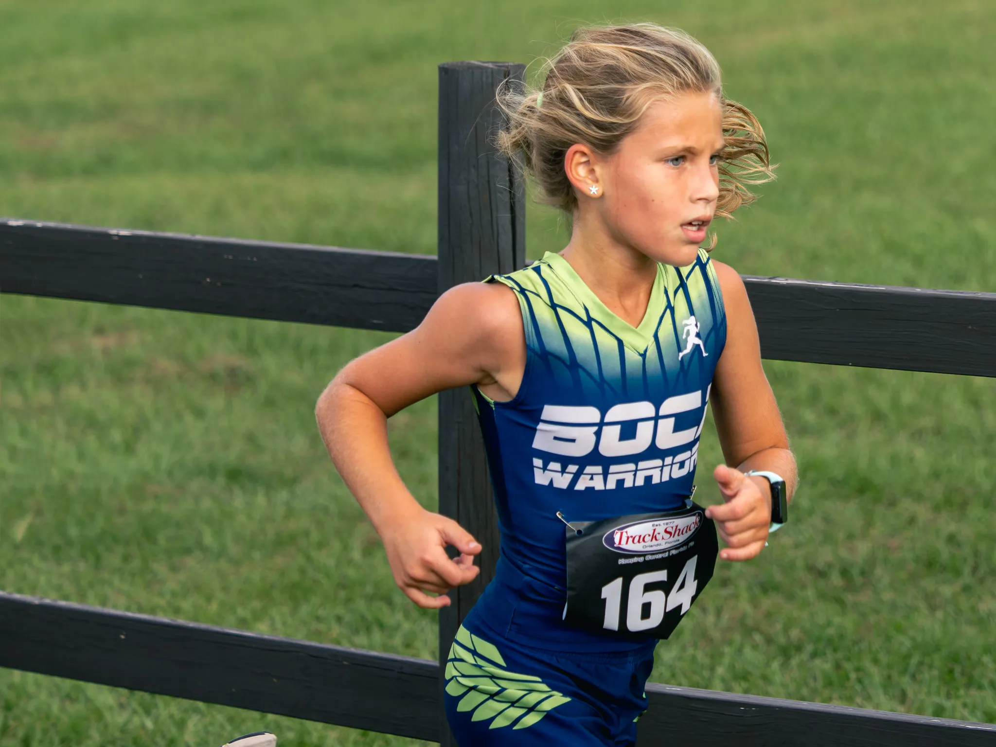 Young blonde girl in blue and green Boca Warriors jersey running cross country past a wooden fence on grass