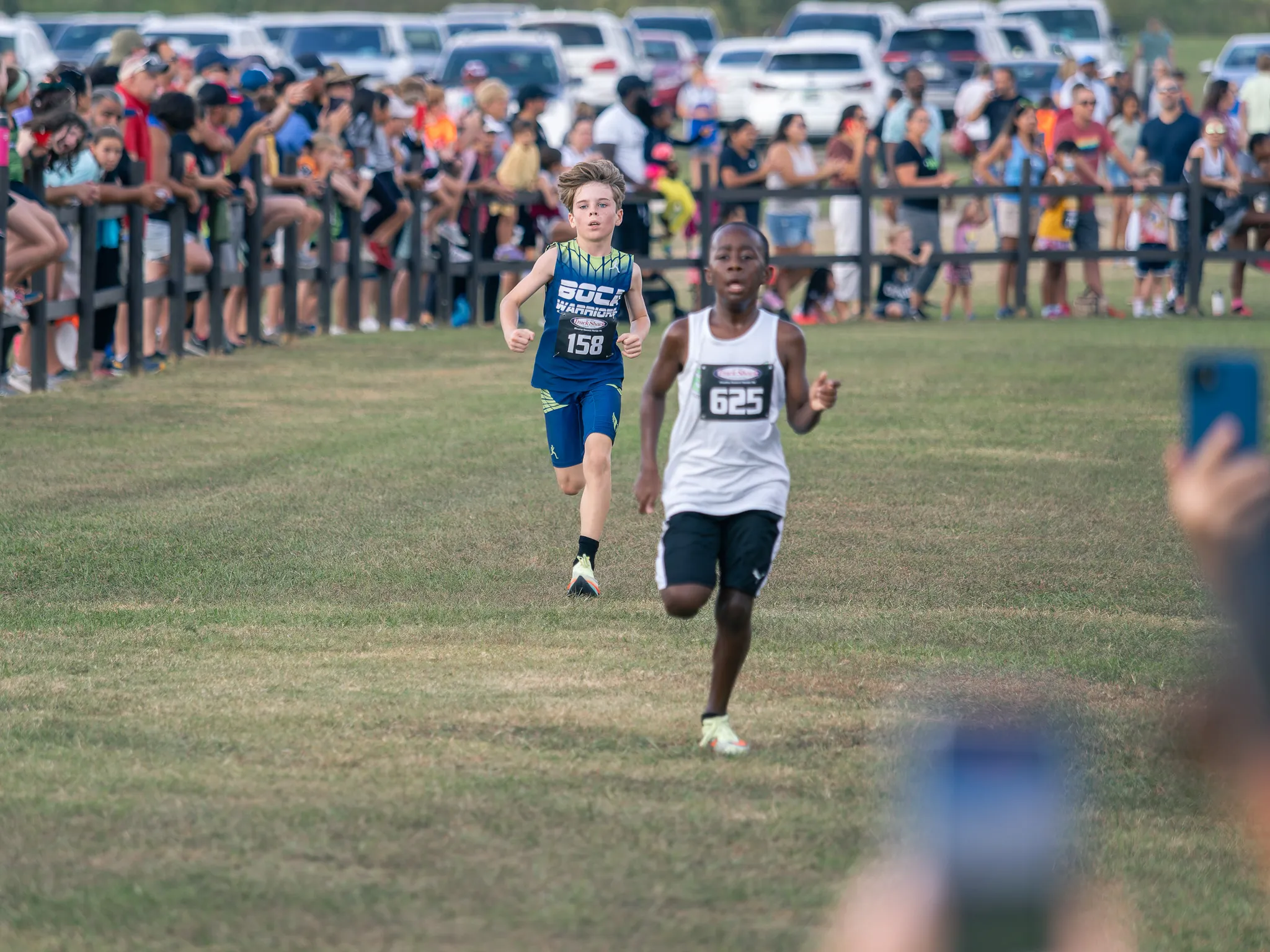 Two youth cross country runners sprinting toward the finish line with spectators watching from behind a fence