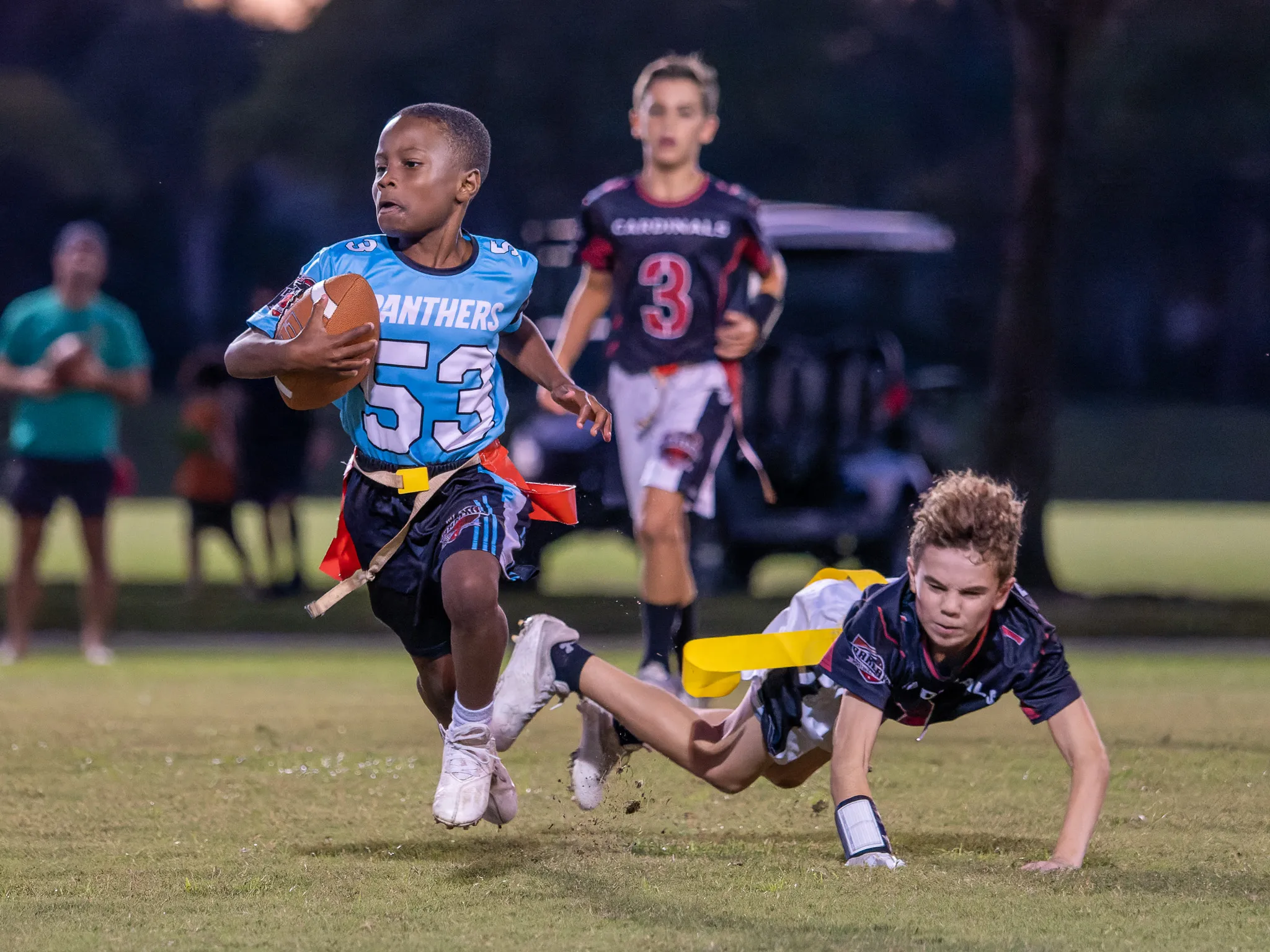 Youth flag football player in blue Panthers jersey number 53 dodging a diving defender under stadium lights