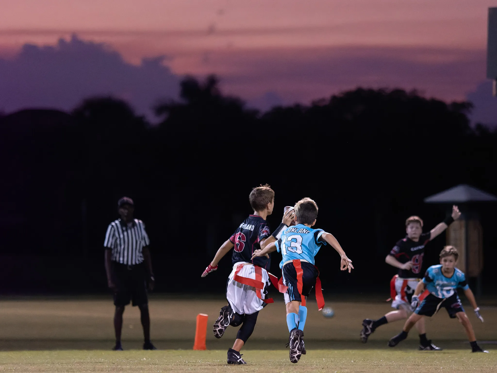 Youth flag football players jumping for a pass under a purple sunset sky with a referee watching nearby
