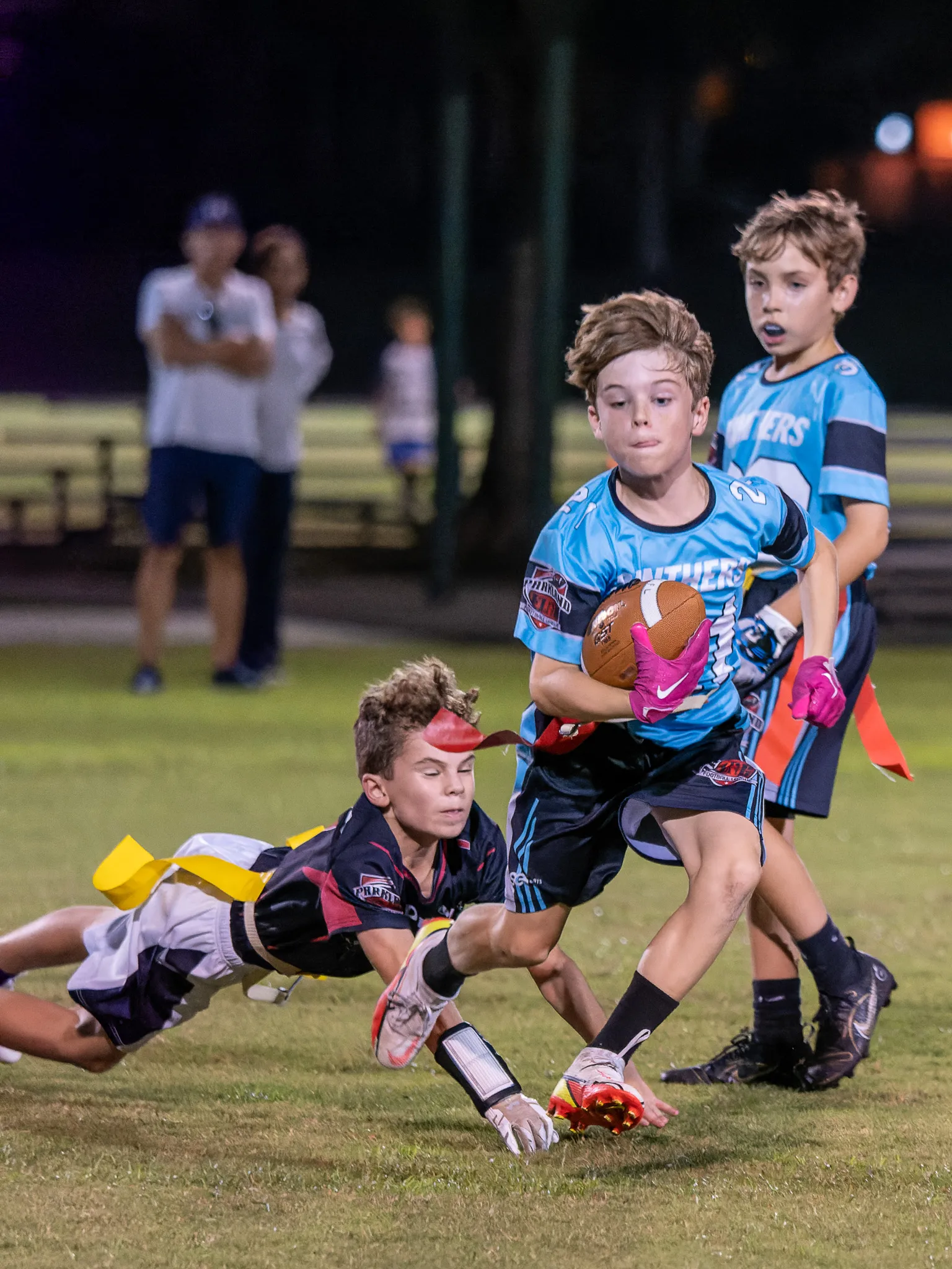 Youth flag football player in blue Panthers jersey carrying the ball while a defender dives for his flags at night