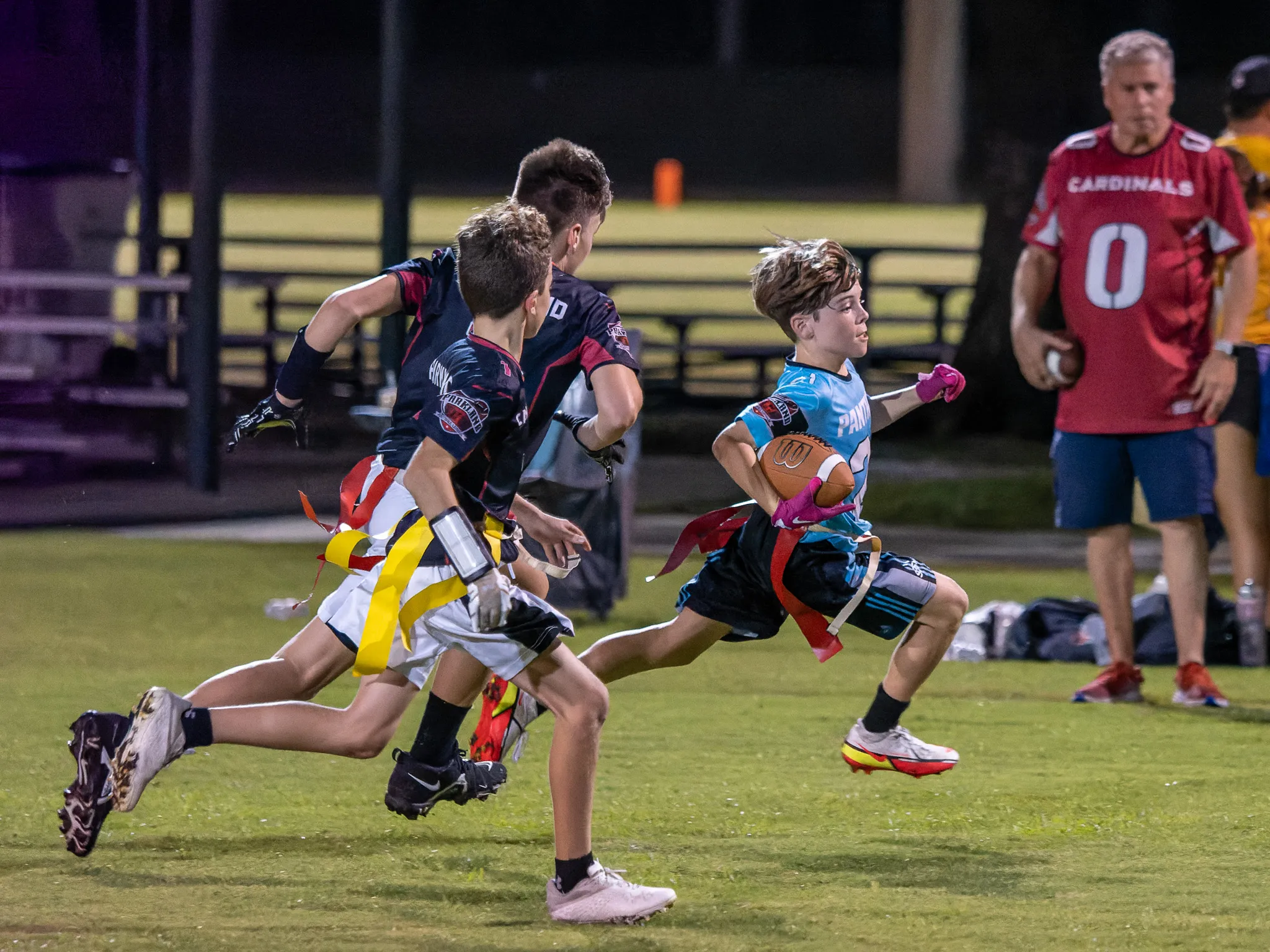 Youth flag football players sprinting on a lit field at night, ball carrier in teal jersey evading two defenders
