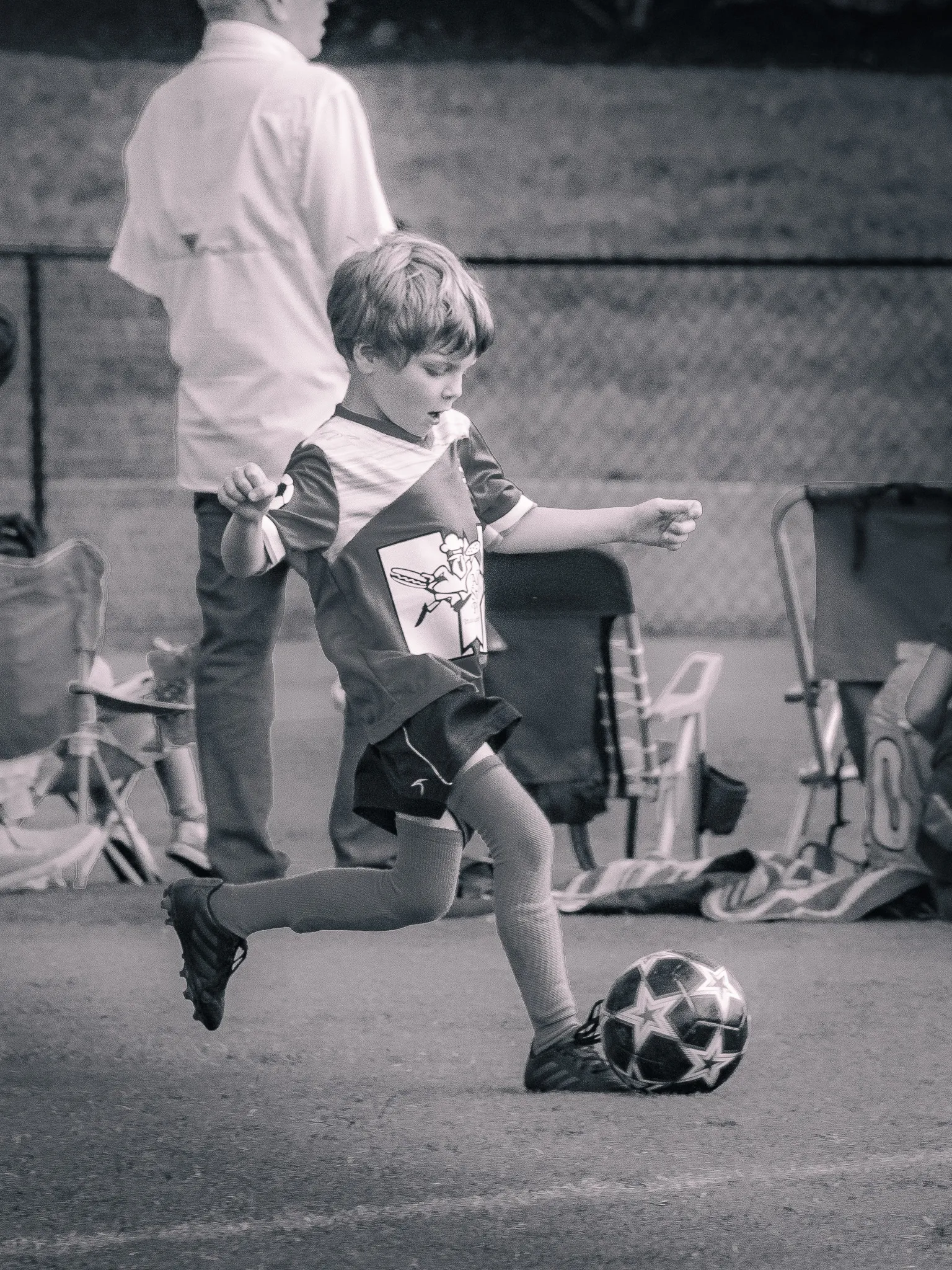 Young boy in soccer uniform kicking a ball mid-stride on a sideline, black and white photo with vintage tone