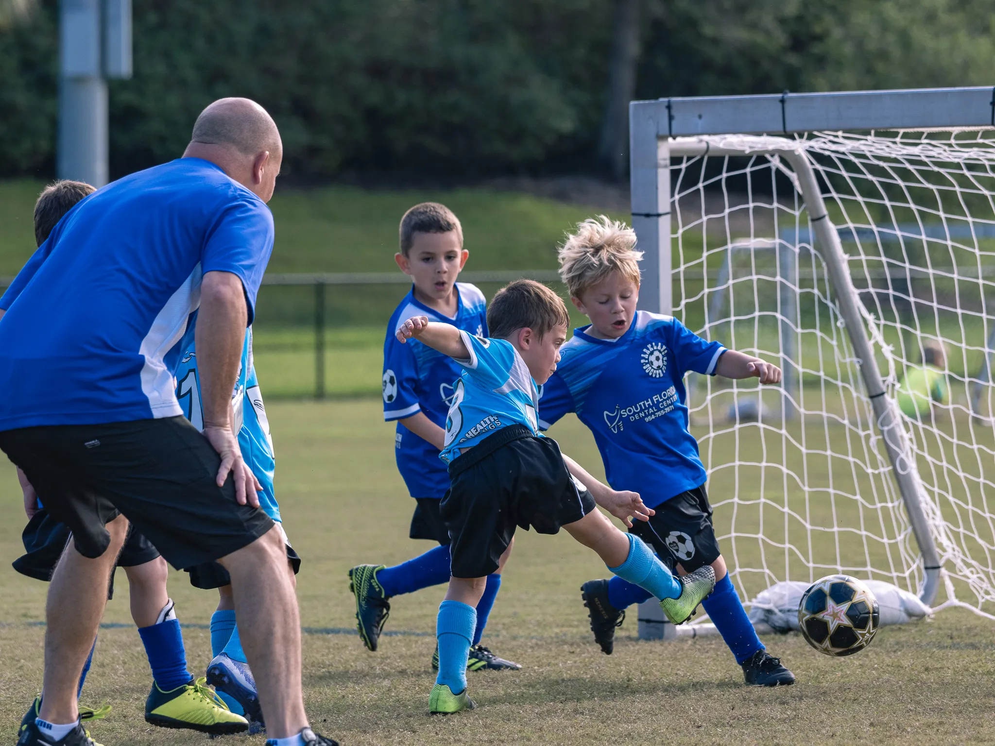 Group of youth soccer players in blue jerseys scrambling at the goal mouth, coach watching from the left side